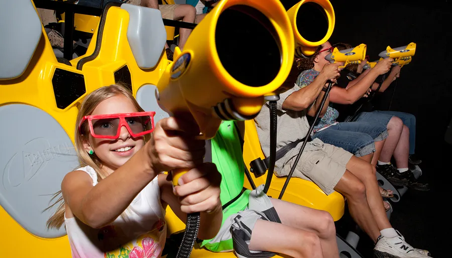 A girl wearing 3D glasses is holding a yellow toy gun while seated in a row with other people, enjoying an interactive amusement ride.