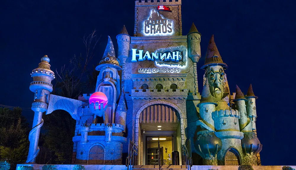 The image depicts a whimsical castle-themed building at night illuminated with lights featuring signs for Castle of Chaos and Hannahs Maze of Mirrors