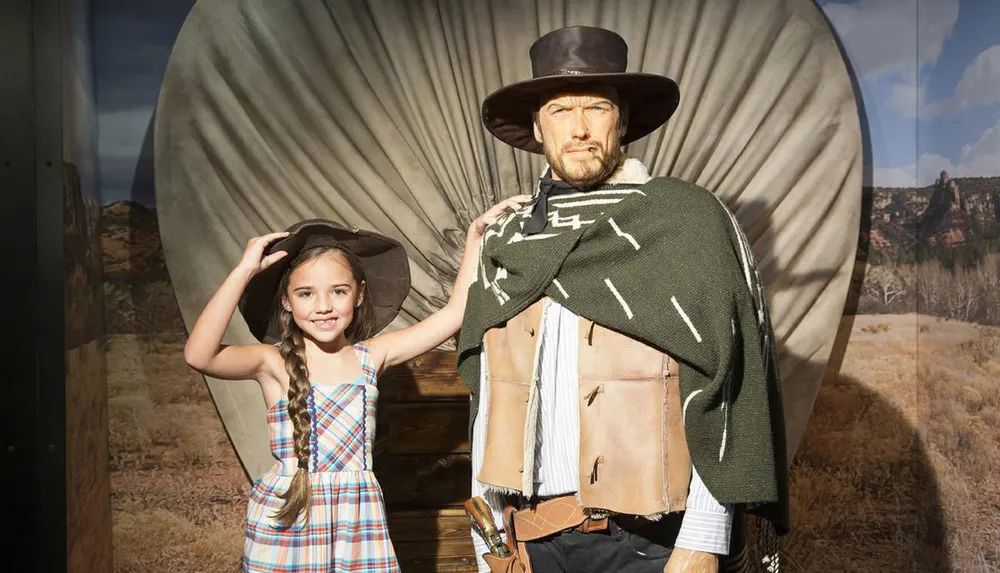 A young girl smiling at the camera poses with a lifelike figure dressed as a cowboy in front of a Western-themed backdrop