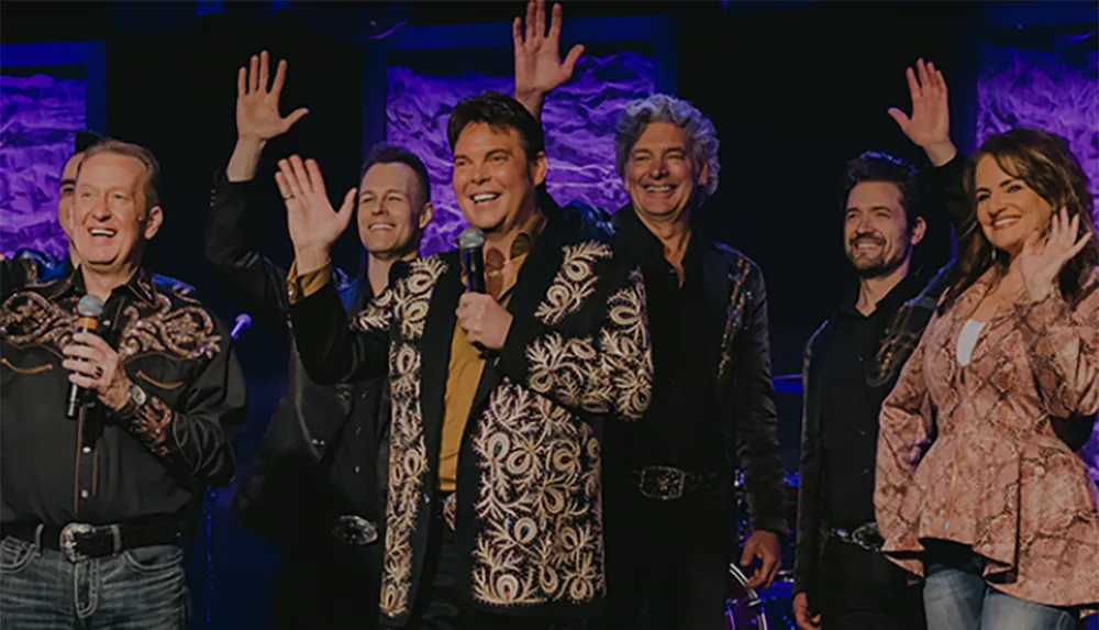 A group of six smiling performers some with raised hands are on stage possibly taking a bow or greeting the audience at an event