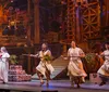 A theater performer dressed in a colorful costume releases a brightly colored parrot towards another actor on stage set against a backdrop suggestive of a village or rustic setting at dusk