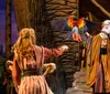 A theater performer dressed in a colorful costume releases a brightly colored parrot towards another actor on stage set against a backdrop suggestive of a village or rustic setting at dusk