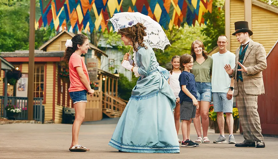 A group of people watches as a young girl interacts with two performers dressed in historical costumes, including a woman in a blue dress with a parasol.