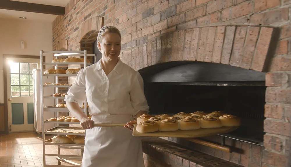 A baker in a white uniform is taking freshly baked cinnamon rolls out of a brick oven