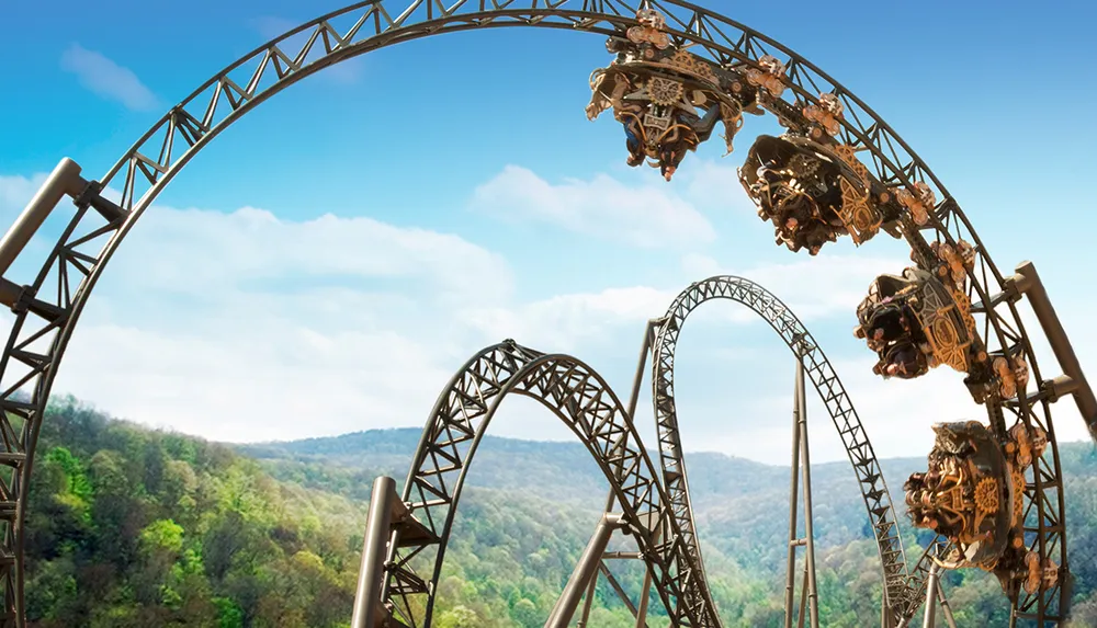 A roller coaster with looping tracks is set against a scenic backdrop of hills and a clear blue sky