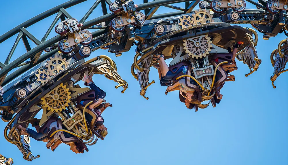 The image shows people riding an inverted roller coaster with intricate mechanical designs against a clear blue sky
