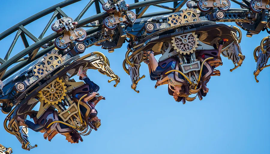 The image shows people riding an inverted roller coaster with intricate mechanical designs against a clear blue sky.