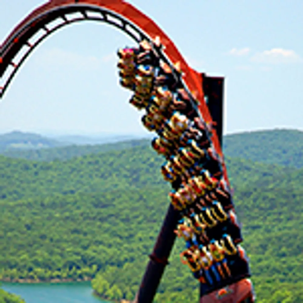 A roller coaster car full of people is descending a steep looping track against a backdrop of lush green hills and a blue sky