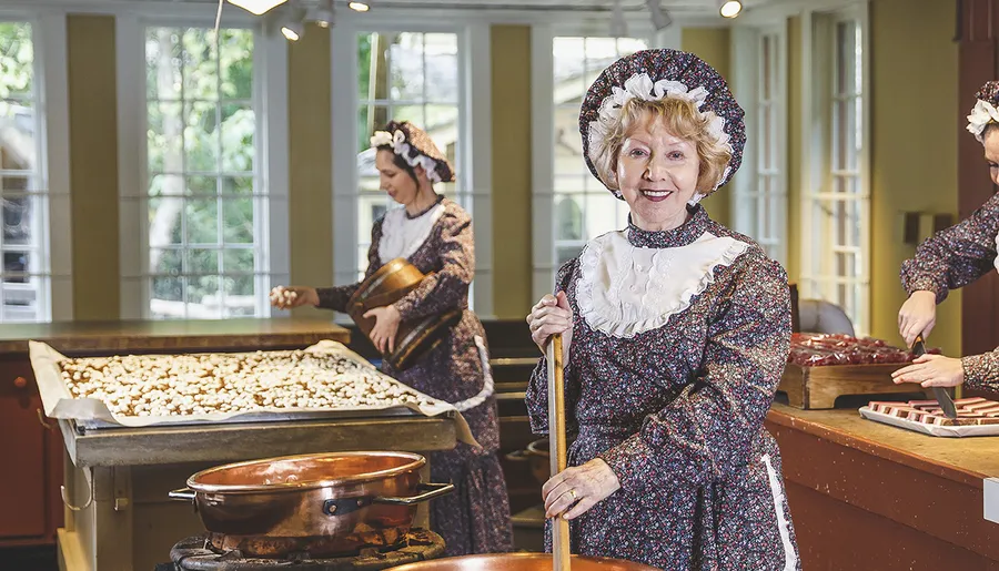 Three women dressed in traditional clothes are making sweets in a brightly lit kitchen.