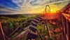 A roller coaster speeds down the track at sunset against a backdrop of lush greenery and a vibrant sky.