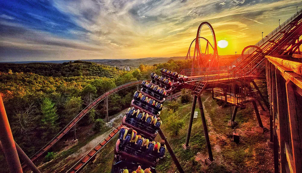 A roller coaster speeds down the track at sunset against a backdrop of lush greenery and a vibrant sky