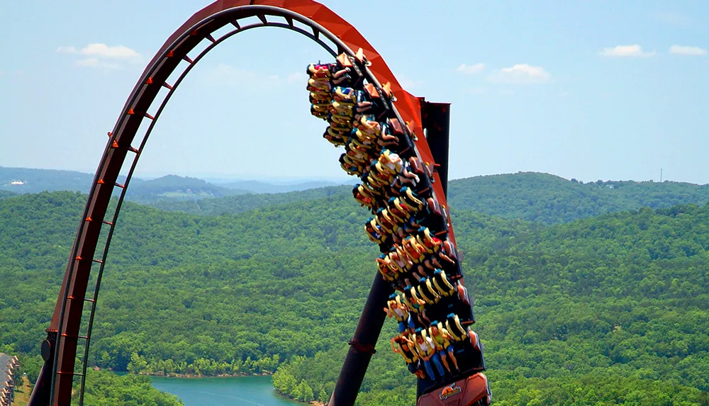 A roller coaster full of riders is ascending a steep loop with a lush green forest in the background