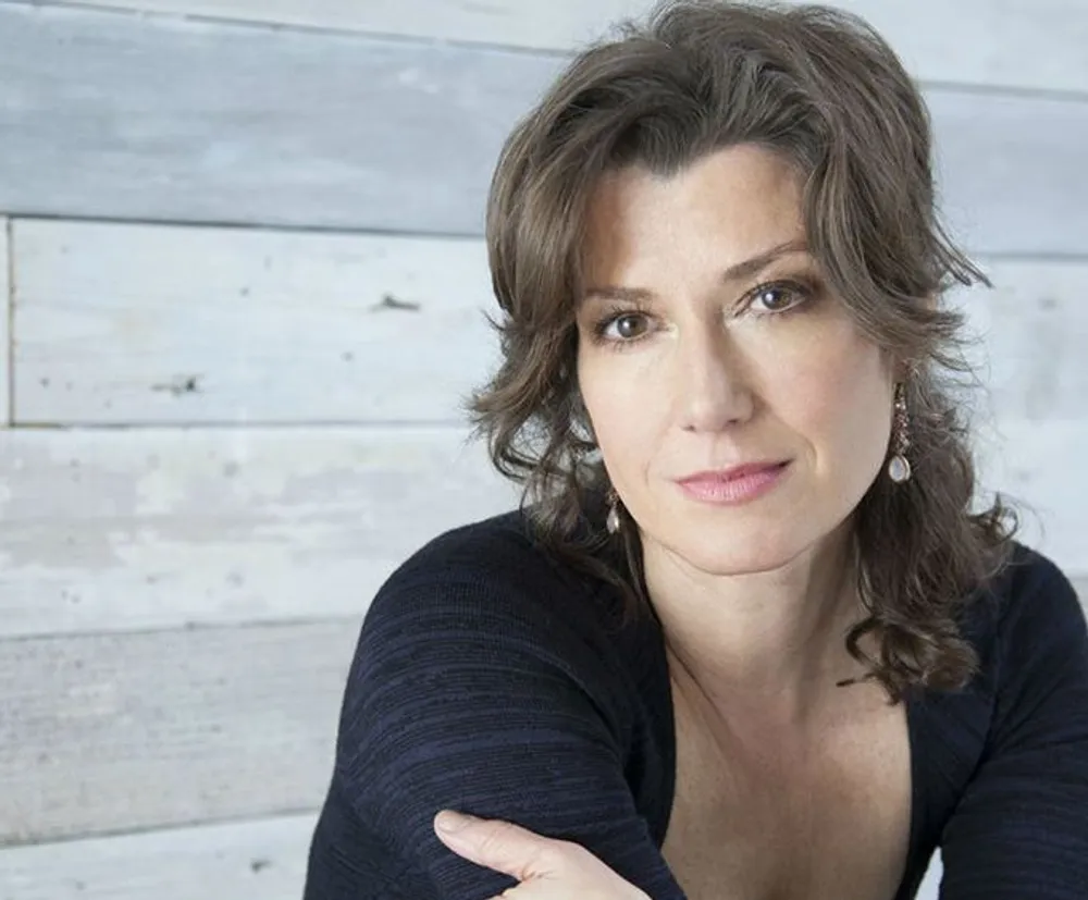 A woman with shoulder-length hair and a subtle smile is posed with one arm crossed in front of her against a background of weathered wooden planks