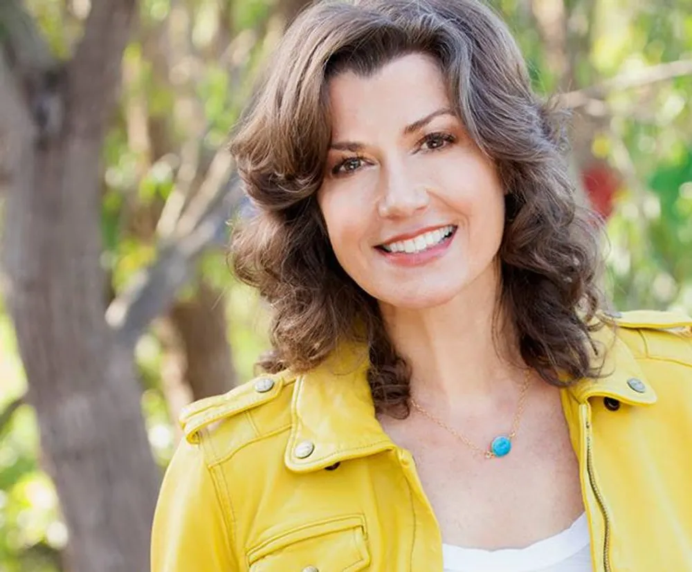 A smiling woman with wavy brown hair wearing a yellow jacket and a white top is posing outdoors with trees in the background