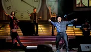 A performer in a cowboy hat is exuberantly singing on stage with musicians and dancers in the background, all set against an oversized clock backdrop.