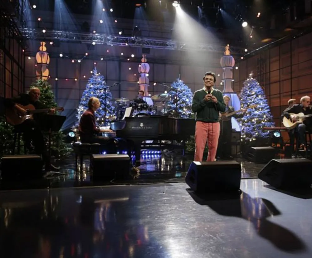 A person is performing on stage with a live band and a festive backdrop of Christmas trees and decorations