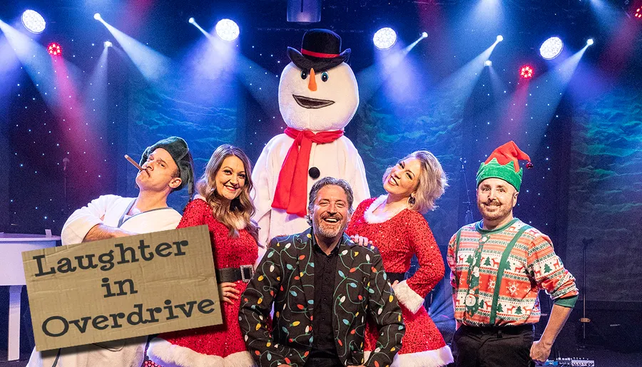 A group of people in festive attire pose with a snowman under stage lights, with a sign reading Laughter in Overdrive.