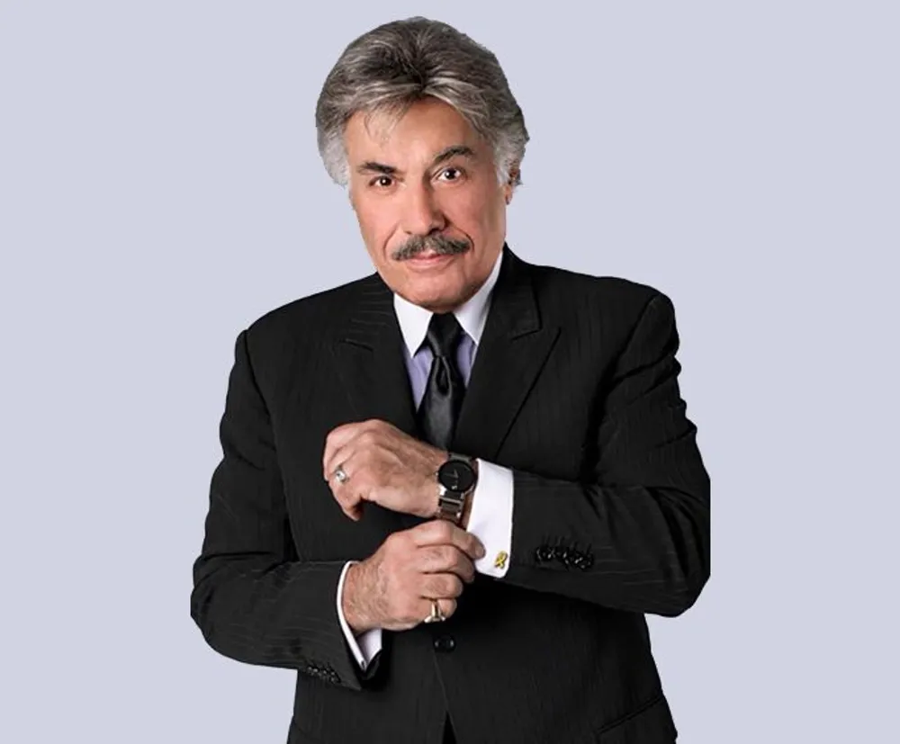 A man with a prominent mustache wearing a suit and watch is posing confidently with one arm crossed over the other against a plain background
