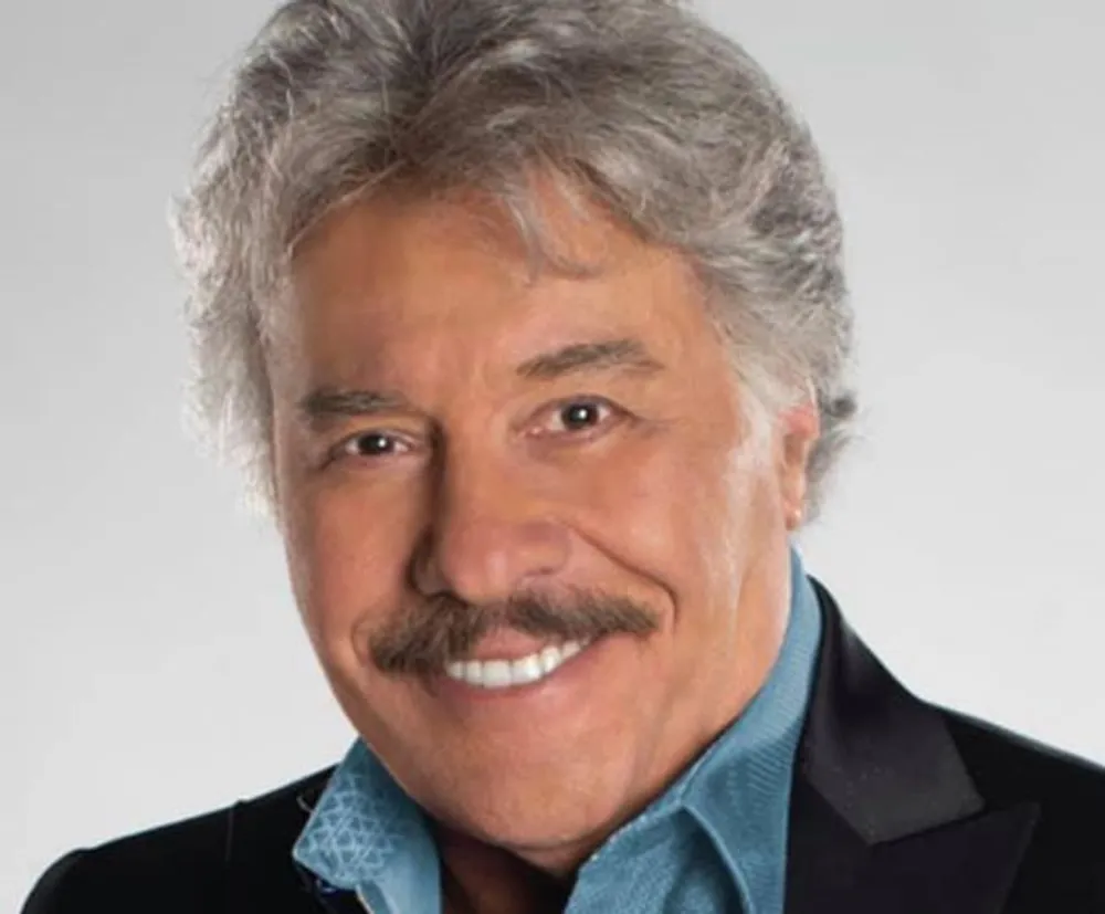 The image shows a smiling man with silver hair and a mustache wearing a black jacket over a blue shirt