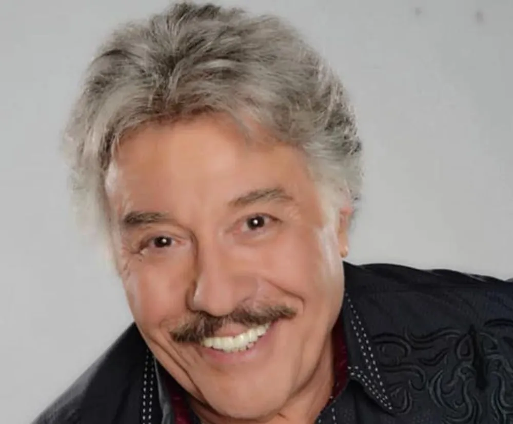 The image shows a smiling man with a mustache salt-and-pepper hair wearing a black shirt with embellished details