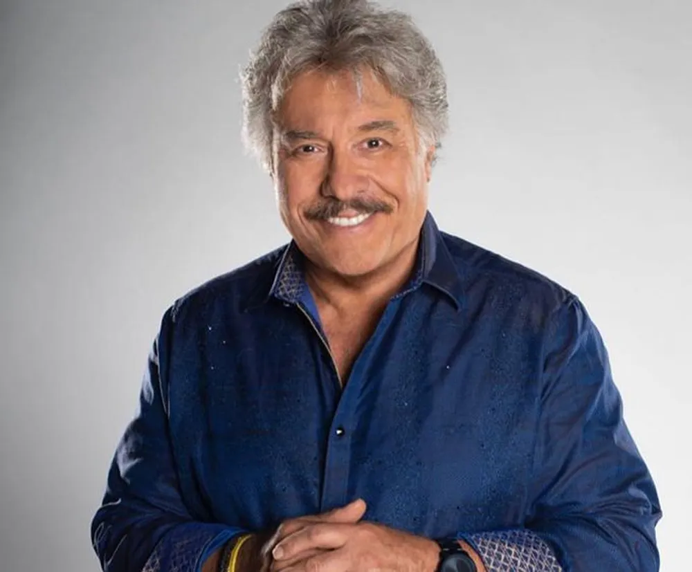 The image features a smiling man with gray hair and a mustache wearing a dark blue shirt with his arms crossed in front of him against a light gray background