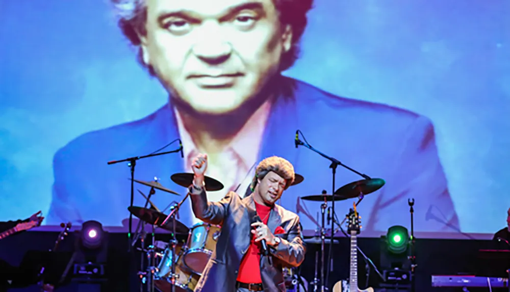 A performer on stage is singing passionately with a large portrait displayed on a screen behind him