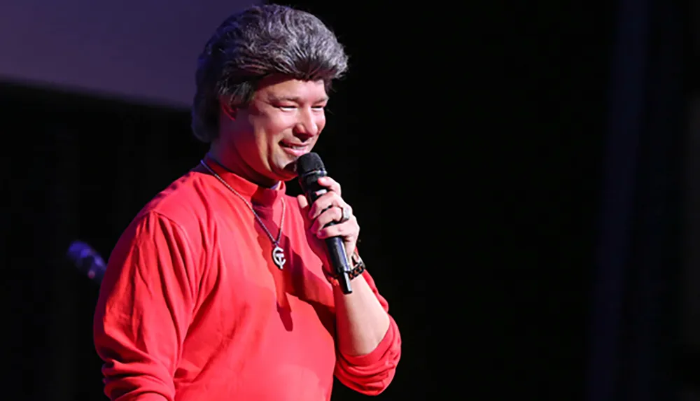 A person is standing on stage wearing a red shirt and speaking into a microphone
