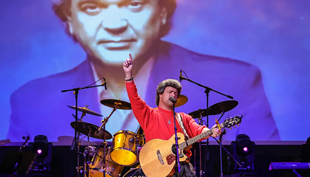 A musician in a red shirt plays guitar and sings on stage with a large black-and-white portrait projected behind him