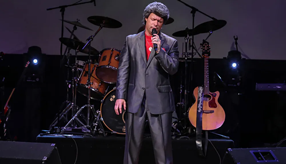 A man in a gray suit is singing on stage in front of a drum set and a guitar