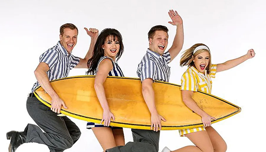Four people are joyfully posing together with a giant yellow surfboard against a white background.