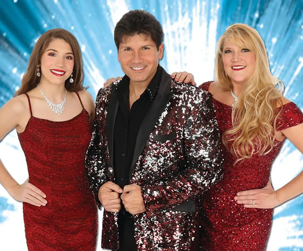 Three smiling performers two women in elegant red sequined dresses and a man in a black shirt with a sequined jacket are posing together with a festive blue background featuring white light bursts