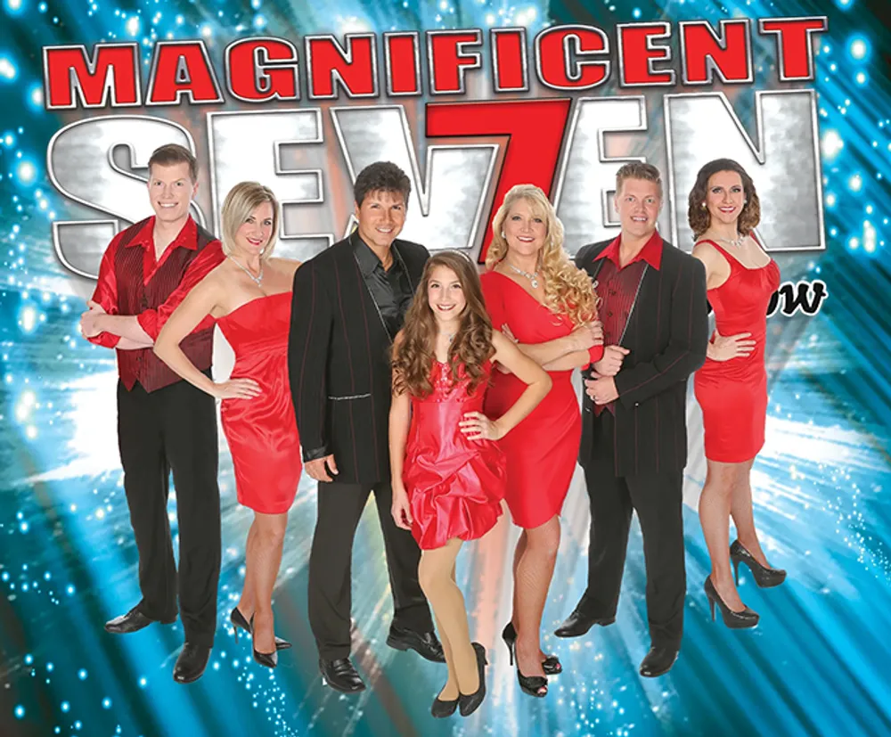 The image shows seven performers dressed in red and black smiling and posing for a promotional photo for their show titled Magnificent Seven