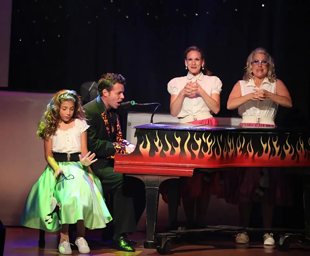 Four individuals are performing on stage with a man playing a piano painted with flames while three others including a young girl enthusiastically participate in what appears to be a lively musical act