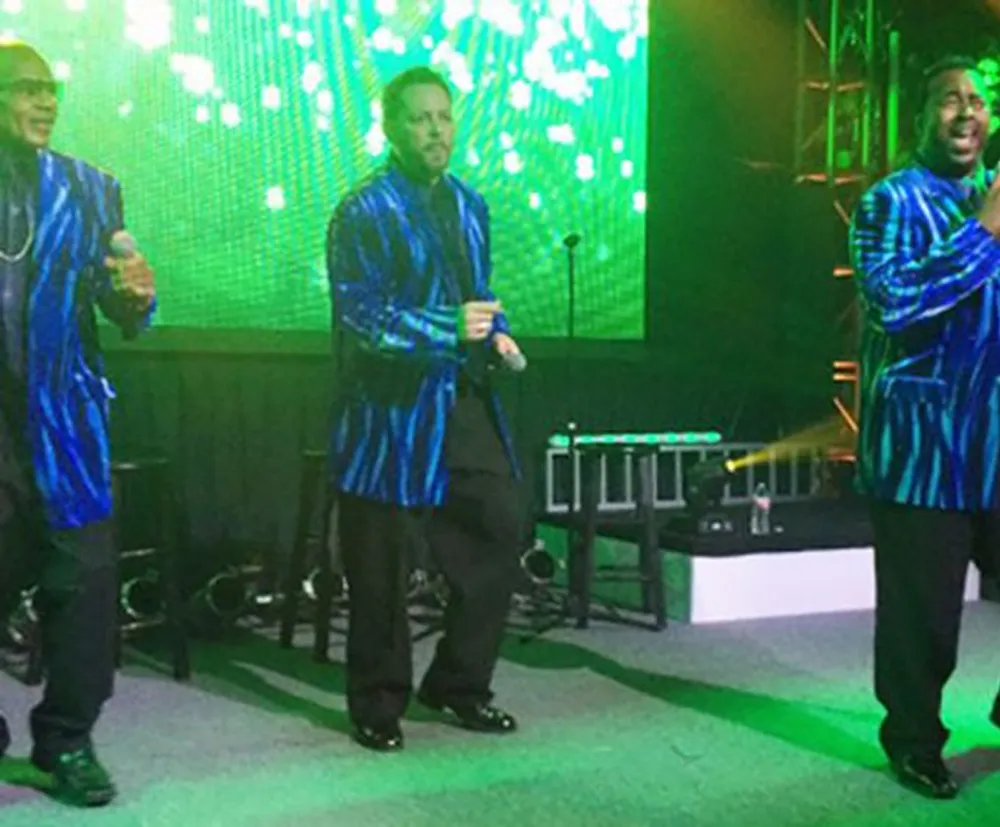 Three men on a stage wear matching blue striped jackets performing with a digital light display in the background
