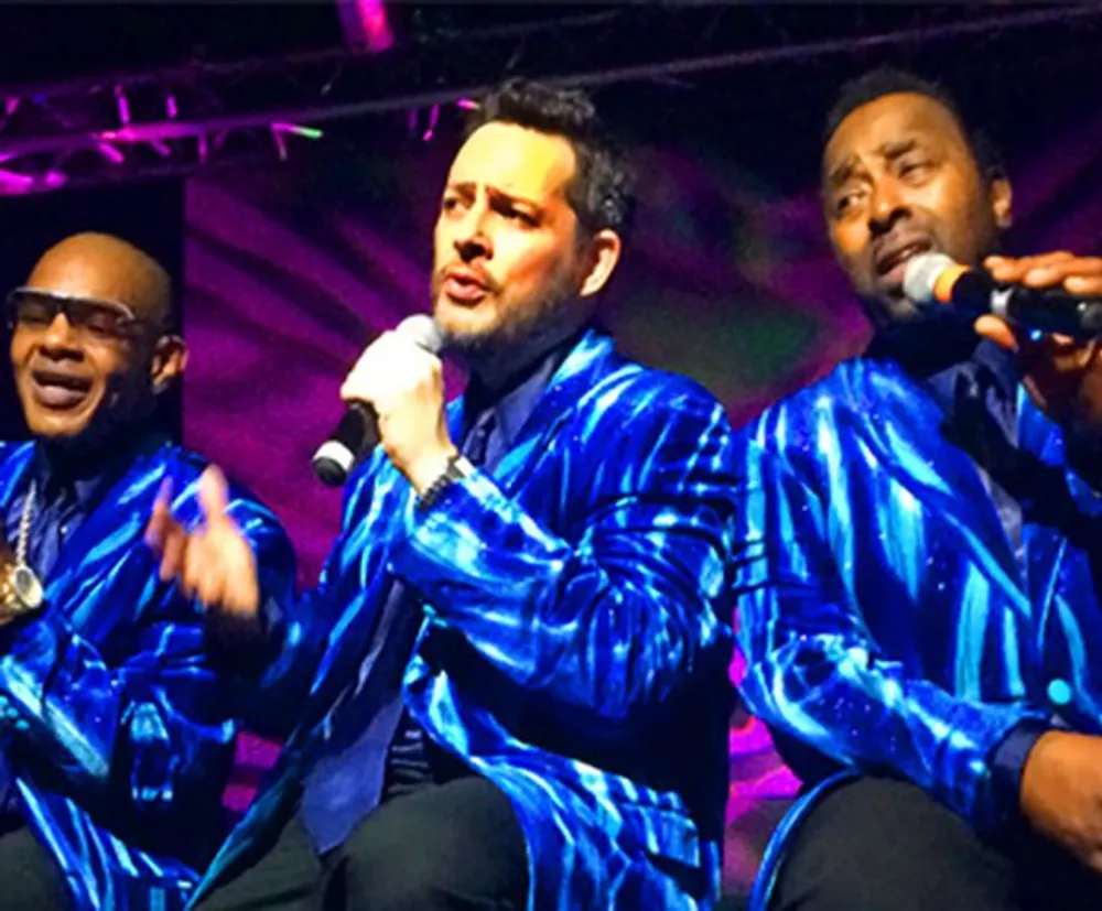 Three men in matching blue shimmering jackets are singing into microphones possibly performing onstage at a music event