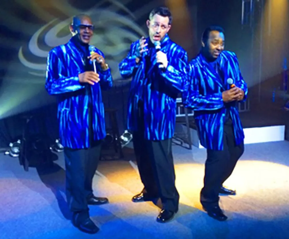 Three singers wearing matching blue patterned jackets are performing with microphones onstage