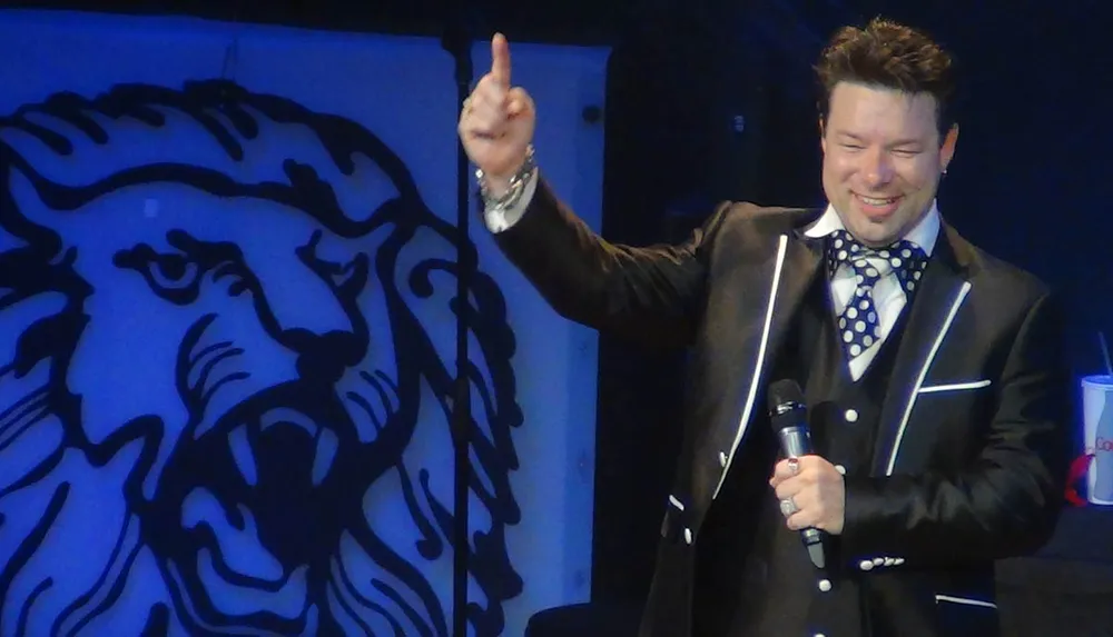 A man in a suit and bow tie is smiling and pointing upwards with one hand while holding a microphone in the other standing in front of a backdrop with a lion design