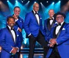 Five men in matching blue tuxedos and bow ties pose confidently on a stage with bright blue lighting and musical equipment in the background