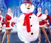 A person in a snowman costume is surrounded by smiling dancers in festive outfits on a winter-themed stage