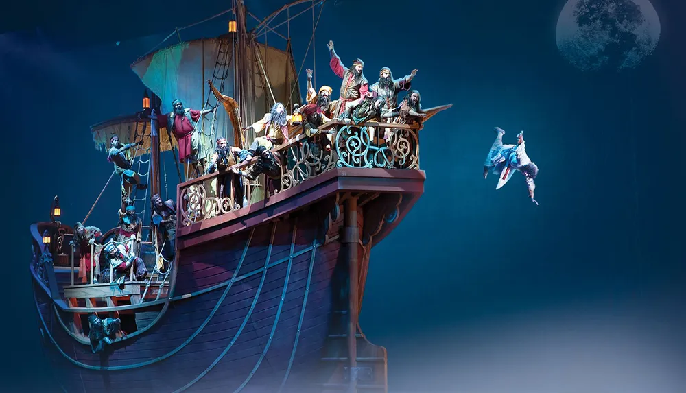 A group of performers dressed as pirates are aboard a dramatically lit stage ship with one character theatrically flying off the ship under a moonlit sky
