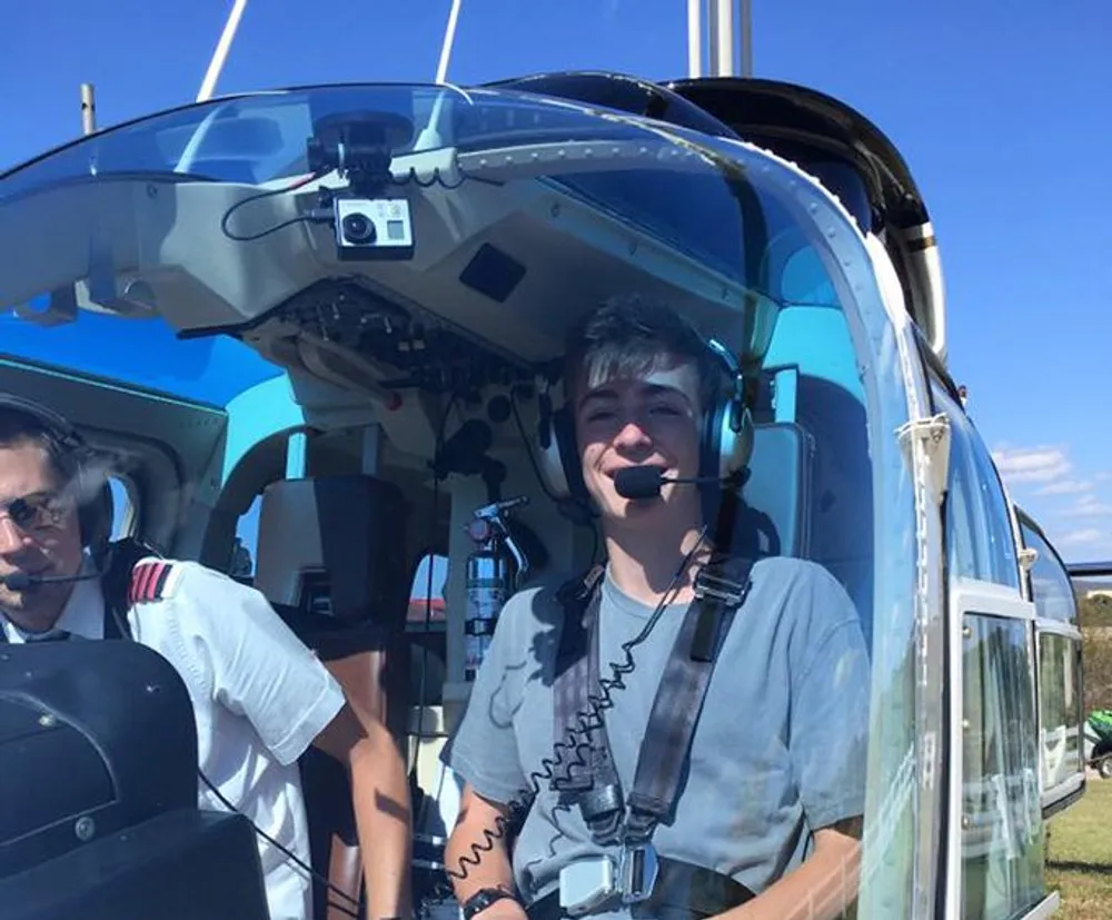 A smiling person with headphones and a microphone is sitting in the front seat of a helicopter with a pilot visible in the periphery