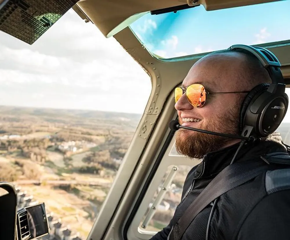 A smiling person wearing sunglasses and a headset is looking out the window of a helicopter observing a landscape from above