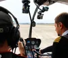 A smiling person wearing sunglasses and a headset is looking out the window of a helicopter observing a landscape from above