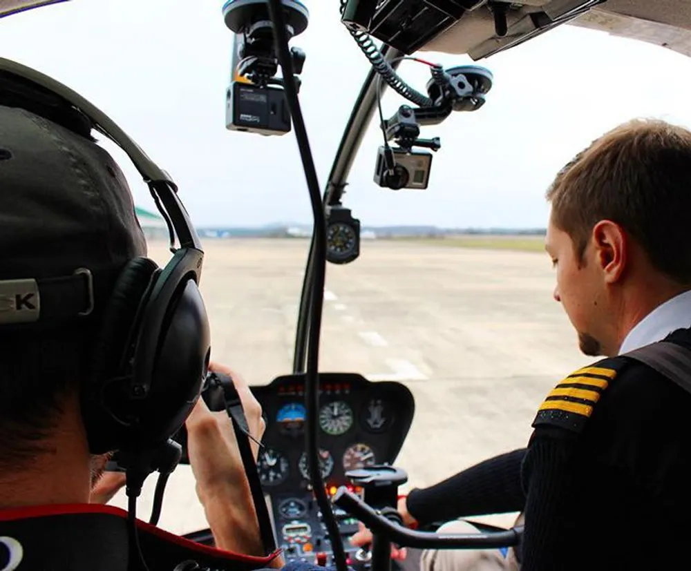 Two pilots are seated in the cockpit of a helicopter preparing for a flight with the runway visible in front of them