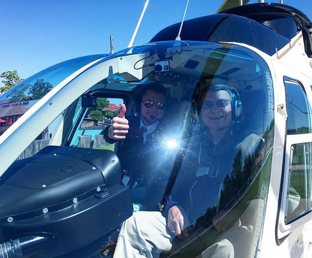 Two individuals are smiling and giving a thumbs-up from the cockpit of a helicopter