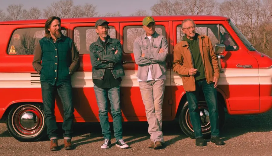 Four men are leaning against a vintage red and white van in a casual outdoor setting.