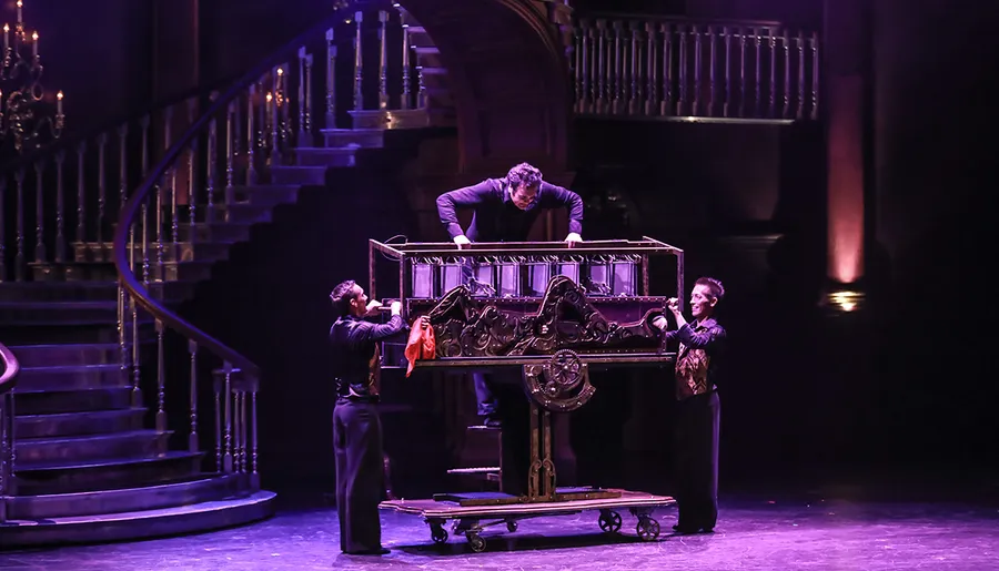 Three performers are seen on stage, with one inside a large ornate box in the midst of a magic trick.