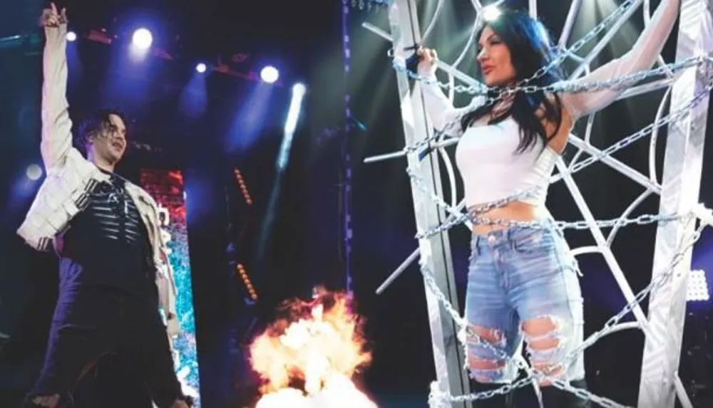 A performer is on stage with one arm raised while a woman stands tied with chains in a metal frame with a fire burning in the background