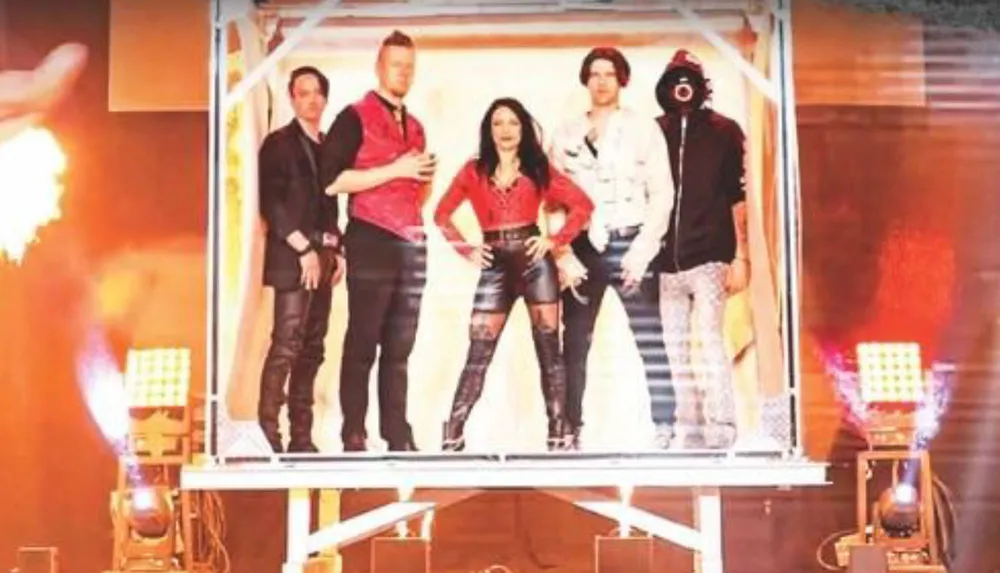 A group of five people dressed in bold stylish outfits poses confidently on a stage with bright lights and flames in the background