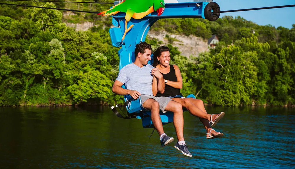 A man and a woman are smiling and enjoying a chairlift ride over a body of water surrounded by lush greenery